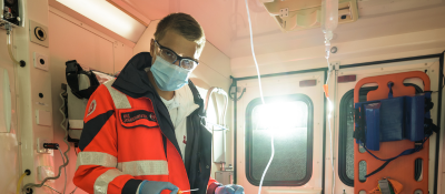 Leon Steinberger trägt Arbeitskleidung sowie eine Gesichtsmaske und eine Schutzbrille. Er steht in einem Rettungsfahrzeug.