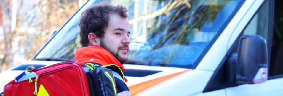 Patrick Jäger steht in Arbeitskleidung vor der Motorhaube eines Rettungsfahrzeugs. Auf dem Rücken trägt er einen Sanitäterrucksack.