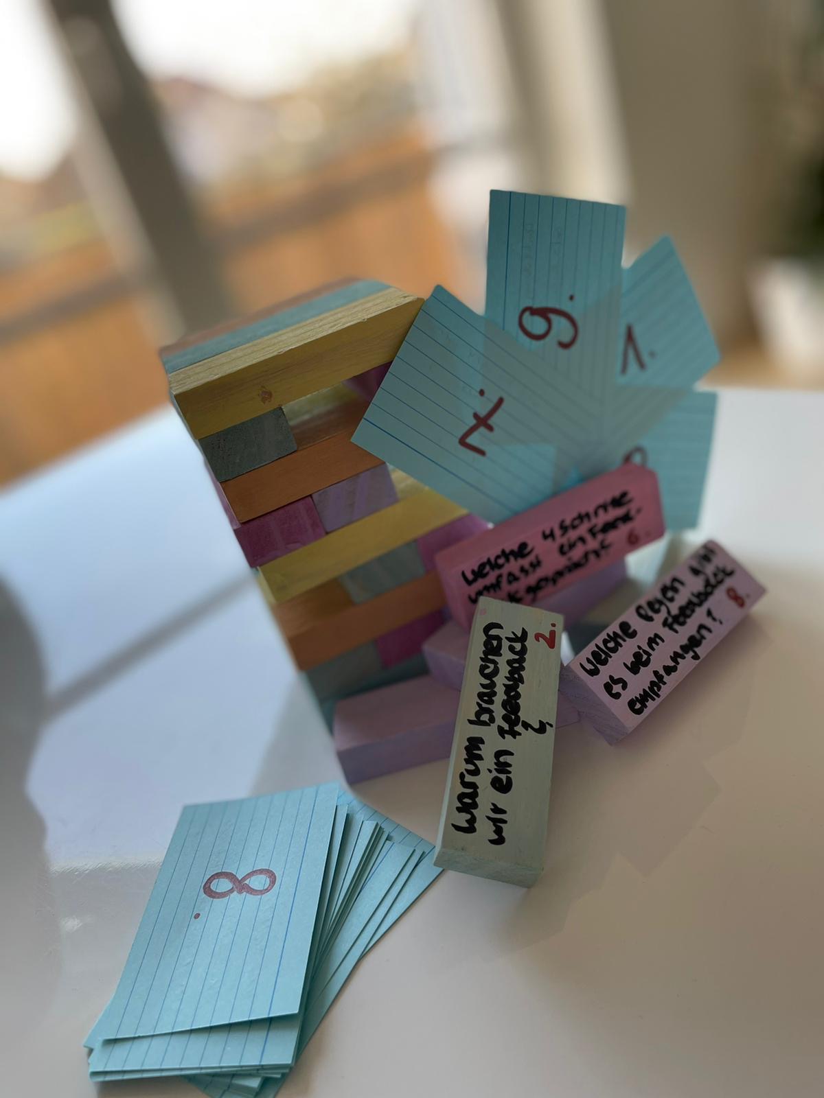 Es ist eine Art Jenga-Spiel zu erkennen oder bunte Holzklötze, auf denen unterschiedliche Fragen draufgeschrieben wurden. Zudem sind Karten, mit dazugehörigen Nummern neben dem Spiel platziert.